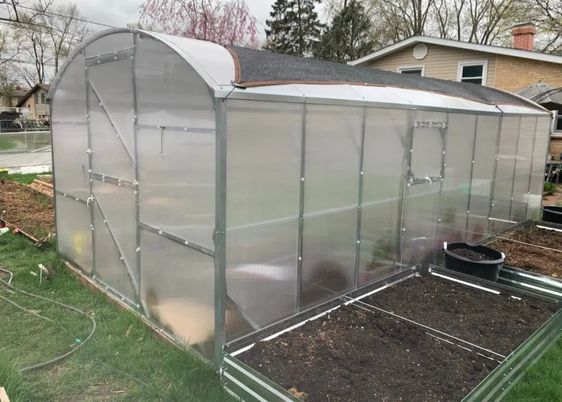 A small arched greenhouse with translucent plastic panels and a gray metal frame, set in a backyard with grass, soil beds, and a house visible in the background.